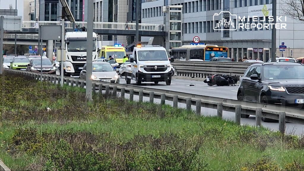 Wielu kierowców zatrzymuje się na ruchliwej drodze, obok leżącego motocykla i pojazdu służb ratunkowych.