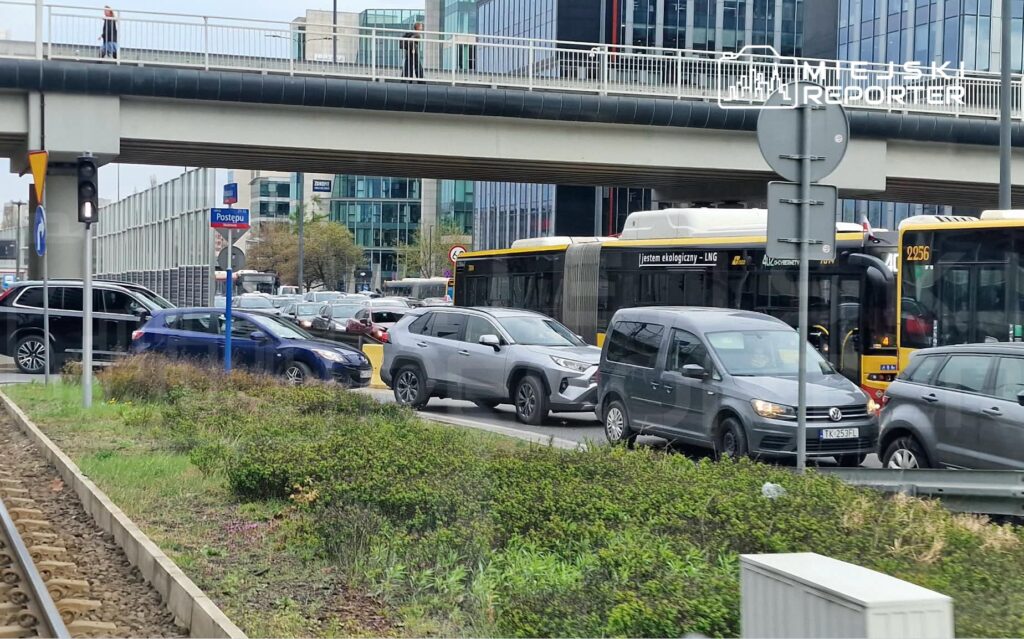 Ruchliwa ulica z wieloma samochodami stojącymi w korku obok przystanku autobusowego "Postępu" oraz nowoczesnych biurowców.