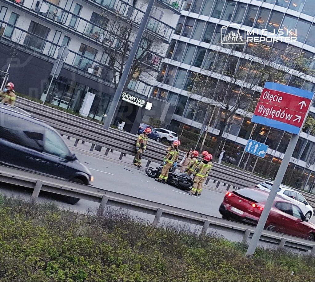 Grupa strażaków w hełmach udziela pomocy przy przewróconym motocyklu na ruchliwej ulicy.