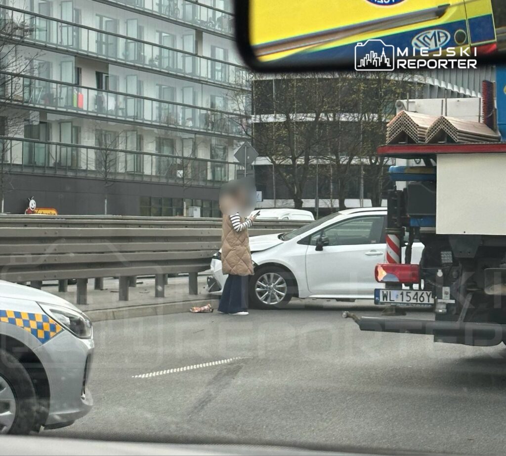 Kobieta w kamizelce z telefonem w ręku stoi obok uszkodzonego samochodu na drodze, w tle nowoczesny budynek.