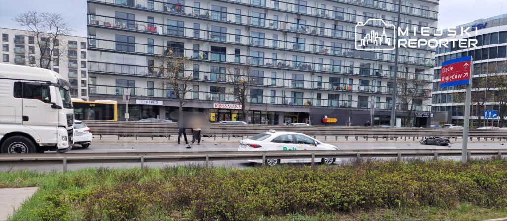 Na jezdni obok białej taksówki stoją dwaj mężczyźni, a w tle widać nowoczesny budynek mieszkalny z balkonami. Na drodze leży przewrócony motocykl.