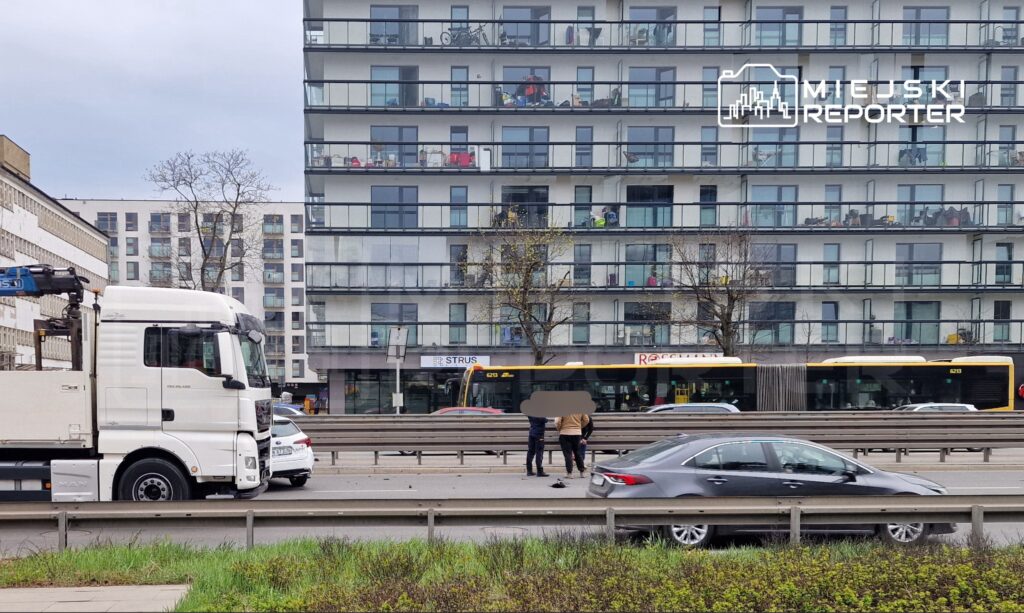 Biały ciężarowy samochód stojący na drodze, obok grupka ludzi rozmawiających, w tle nowoczesny budynek mieszkalny.