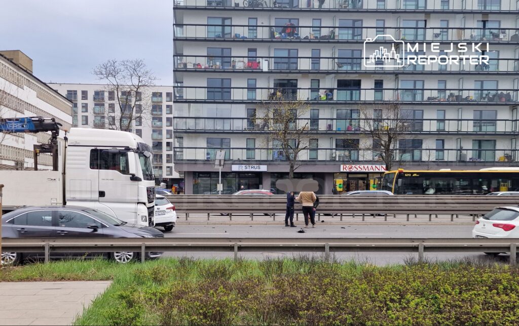 Kierowca w białym samochodzie ciężarowym zatrzymuje się obok dwóch mężczyzn na jezdni, w tle nowoczesne budynki.