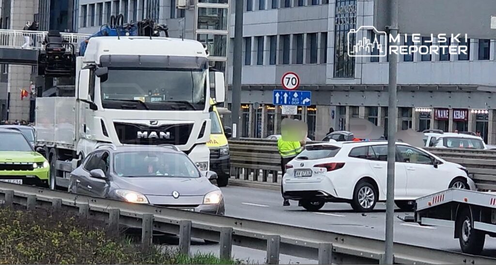Kierowca w żółtej kamizelce kieruje ruchem na zatłoczonej ulicy, obok stoi ciężarówka i kilka osobowych samochodów.