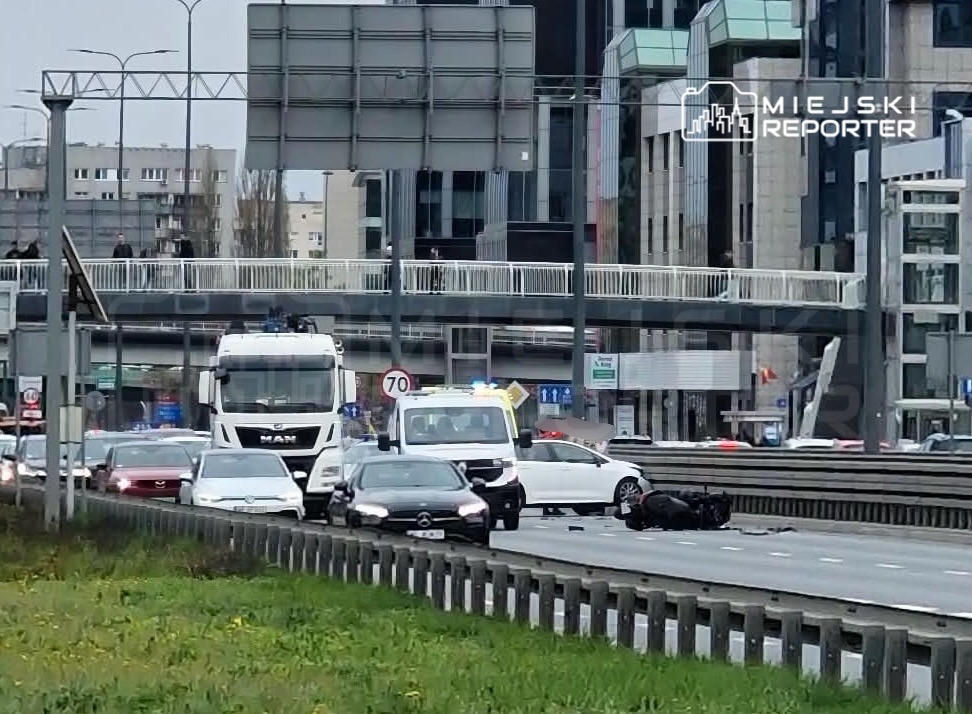 Na jezdni leży przewrócony motocykl, a wokół stoją zablokowane samochody. W tle widać nowoczesne budynki i przejście dla pieszych.