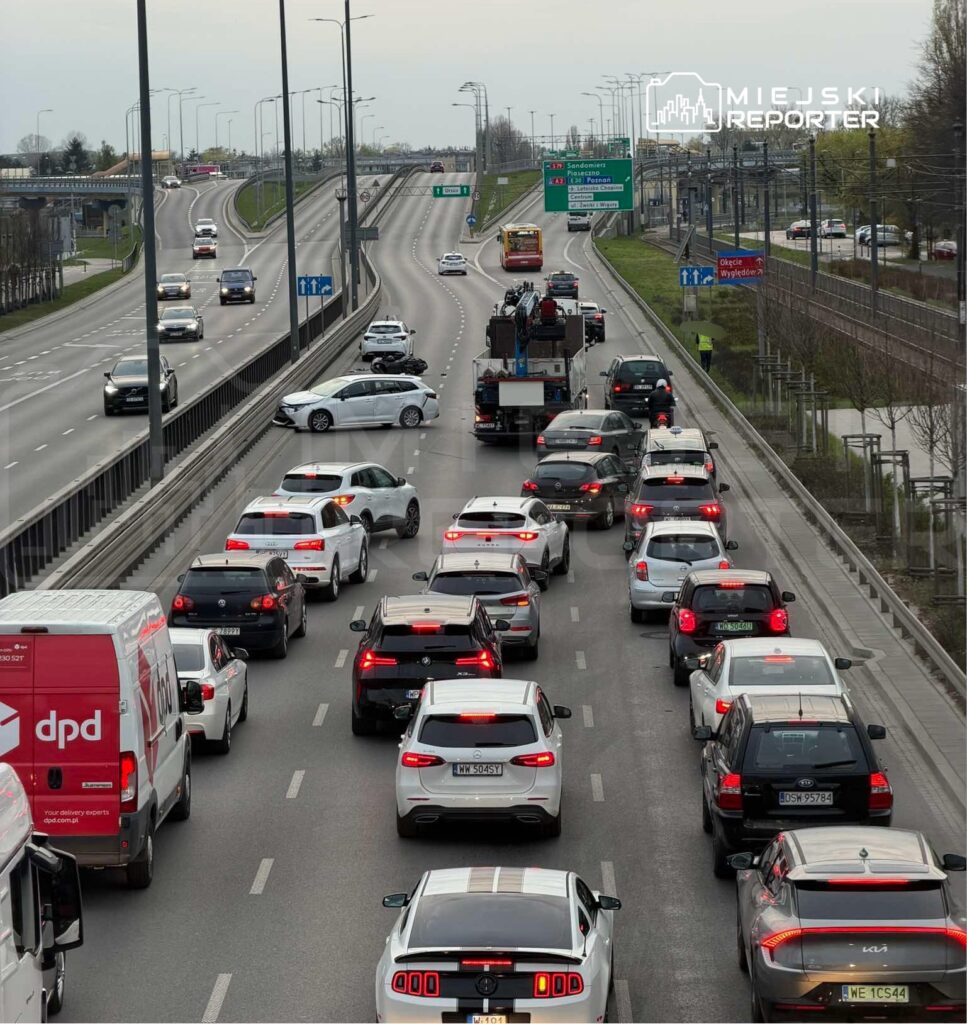 Zatory drogowe na wielopasmowej trasie, z widocznymi samochodami osobowymi i dostawczymi, w tle znaki drogowe.