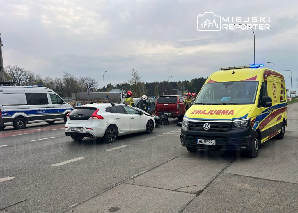 Na skrzyżowaniu zatrzymane pojazdy, w tym białe auto, obok strażacy i karetka pogotowia.