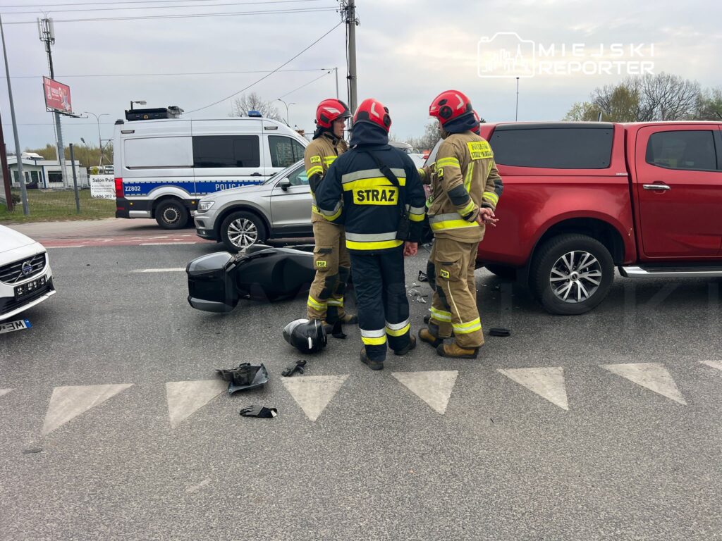 Czterech strażaków w mundurach omawia sytuację obok przewróconego skutera na jezdni. W tle widać policyjny radiowóz.