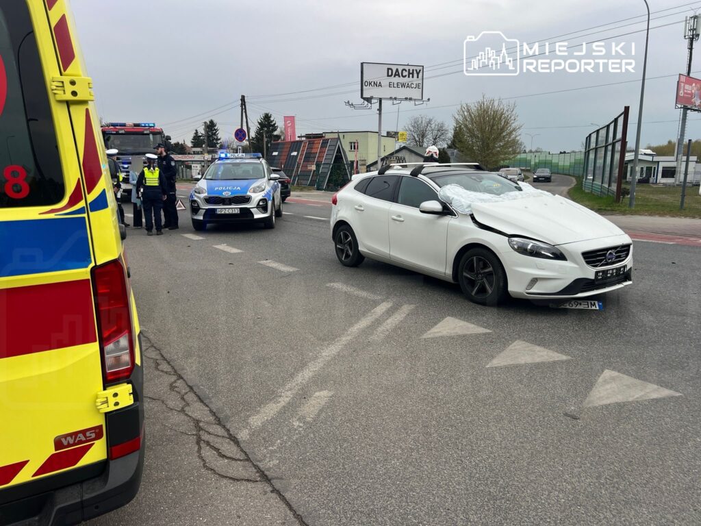 Biały samochód osobowy z uszkodzoną maską stoi na drodze, obok grupy policjantów i karetki pogotowia.