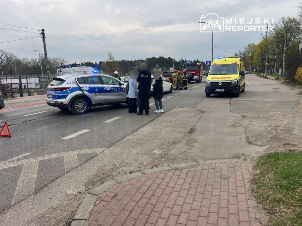 Na drodze zatrzymane pojazdy, policjanci rozmawiają z osobami na miejscu wypadku, w tle karetka i strażacy.
