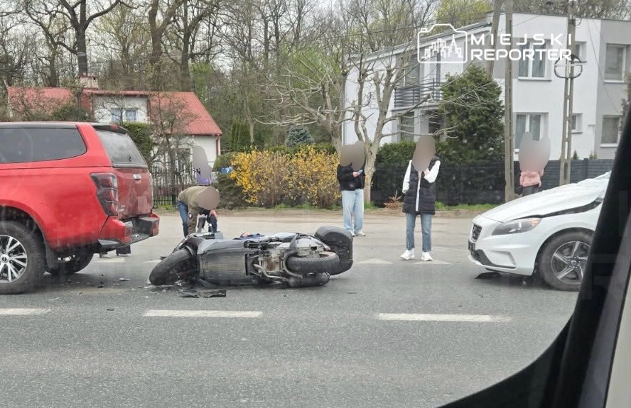 Na jezdni leży przewrócony motocykl obok czerwoną SUV-a, a przechodnie obserwują zdarzenie.