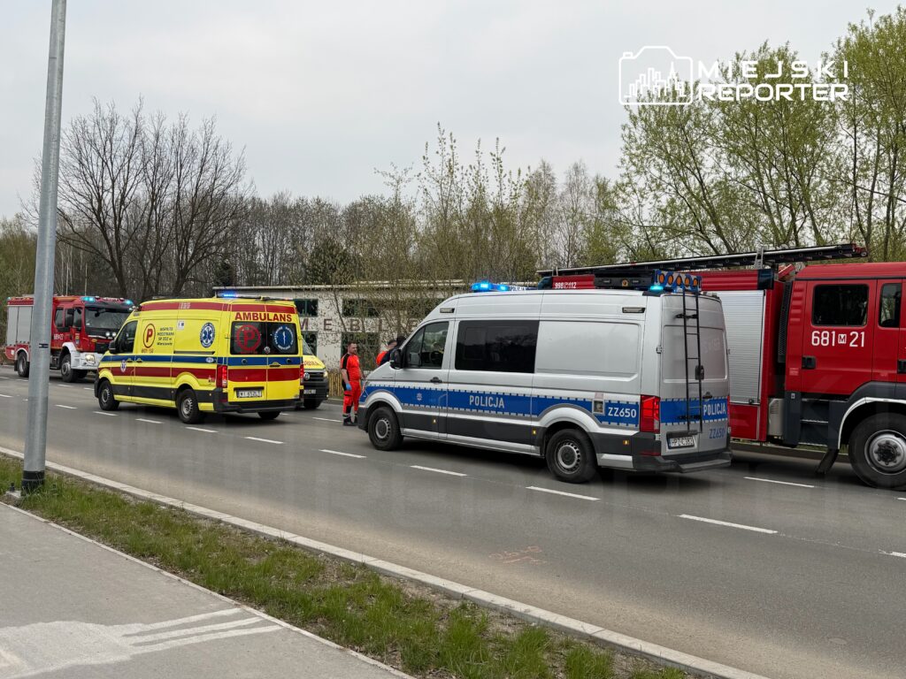 Na drodze zatrzymane są pojazdy służb ratunkowych, w tym karetka, radiowóz i wóz strażacki, obok pracownicy w odzieży ochronnej.