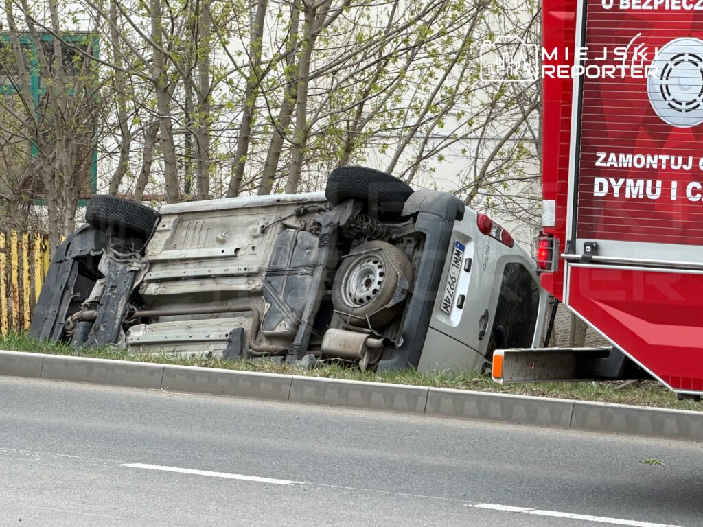 Przewrócony samochód dostawczy leży na boku przy drodze, obok czerwony wóz strażacki z widocznym napisem o bezpieczeństwie.