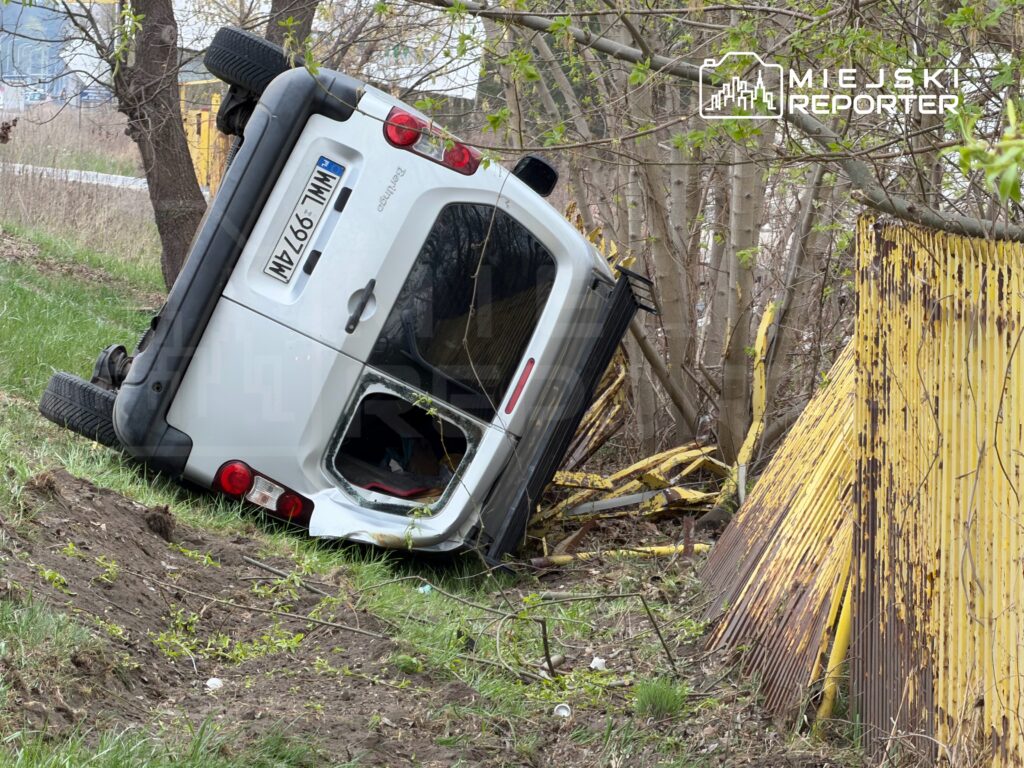 Złomowany biały samochód osobowy leży na boku wśród drzew obok zardzewiałego, żółtego ogrodzenia.