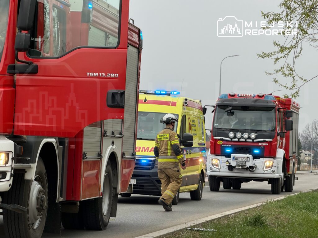 Strażak w mundurze podchodzi do wozu strażackiego na drodze, obok zatrzymany ambulans.