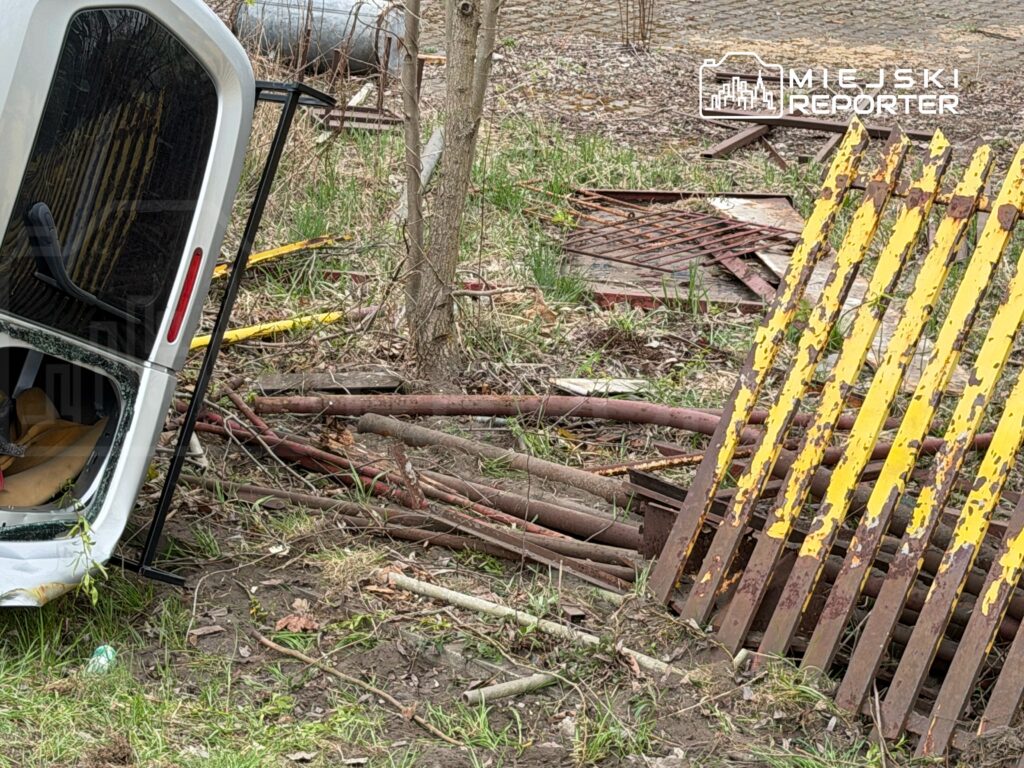 Zniszczona przyczepa leży w zaroślach, otoczona metalowymi rurami i starymi, pomalowanymi na żółto deskami.