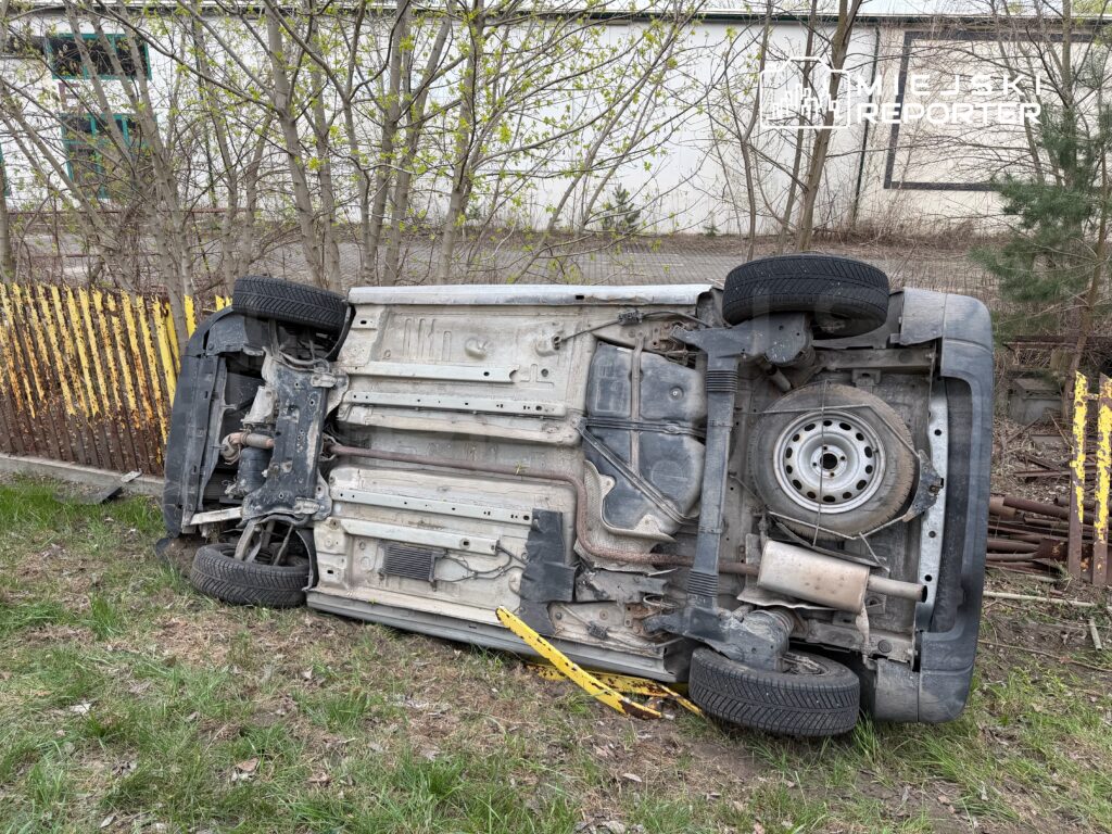 Przewrócone na dach samochód leży na trawie w pobliżu ogrodzenia, w tle widoczne drzewa i budynek.