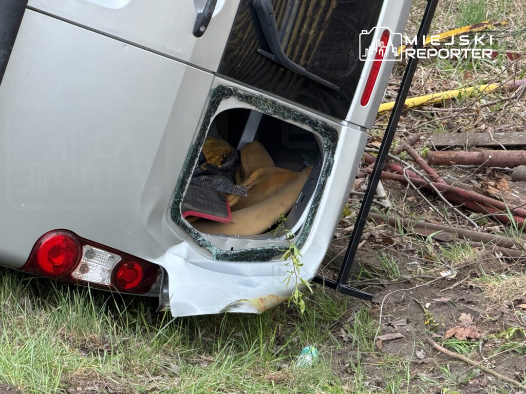 Przewrócony pojazd dostawczy z uszkodzoną tylną częścią leżący na trawie w otoczeniu gałęzi i zarośli.