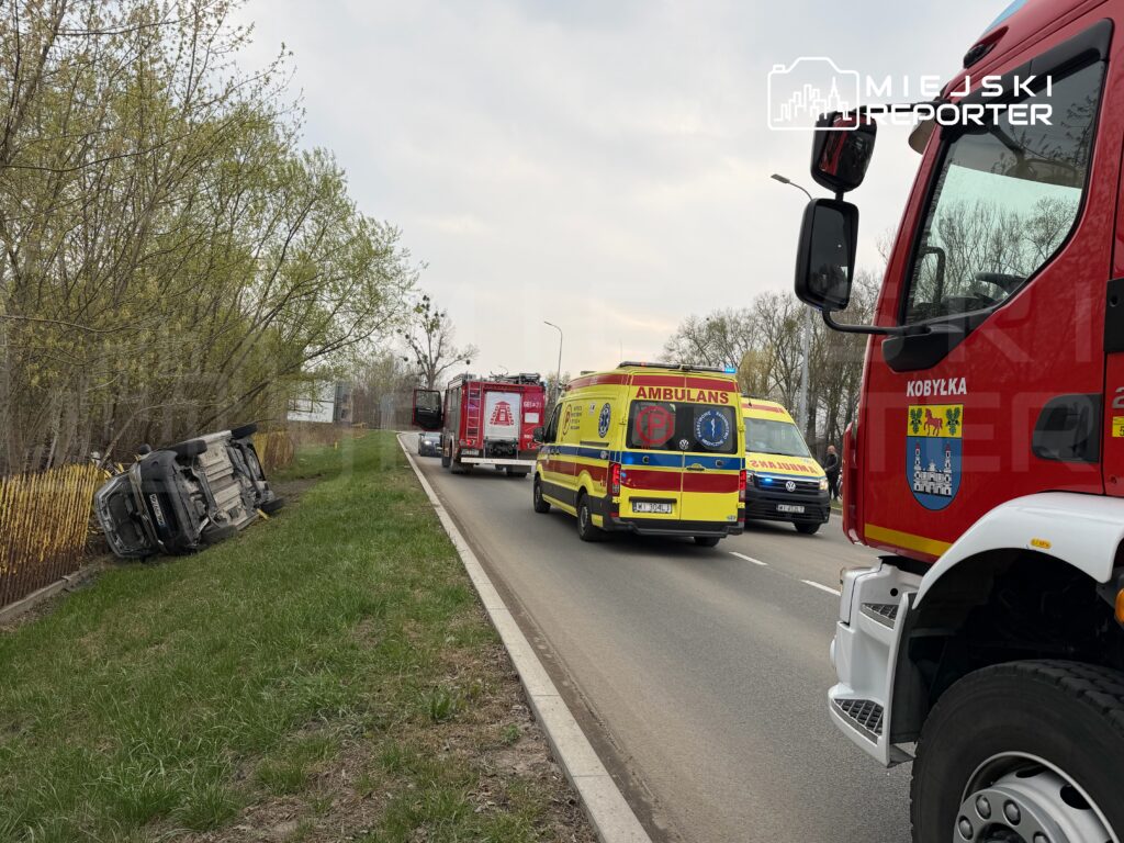 Na poboczu drogi leży przewrócony samochód osobowy, obok zatrzymane pojazdy straży pożarnej i karetka.