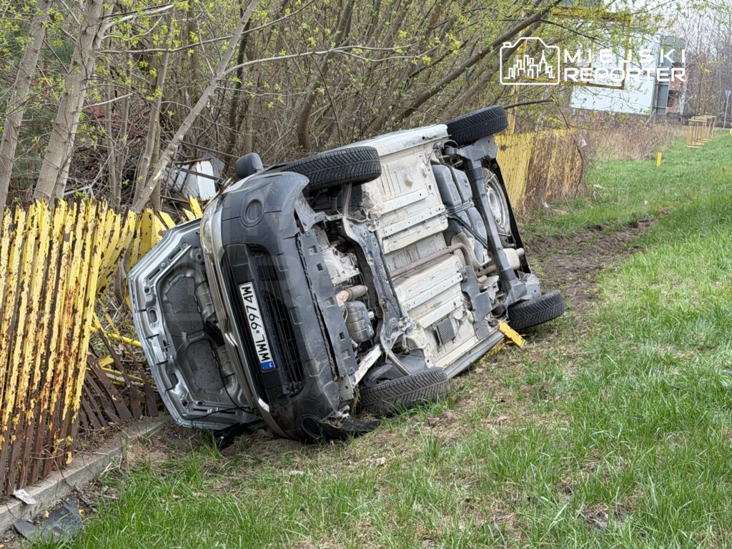 Samochód leżący na dachu w zaroślach obok żółtej, zardzewiałej ogrodzenia.