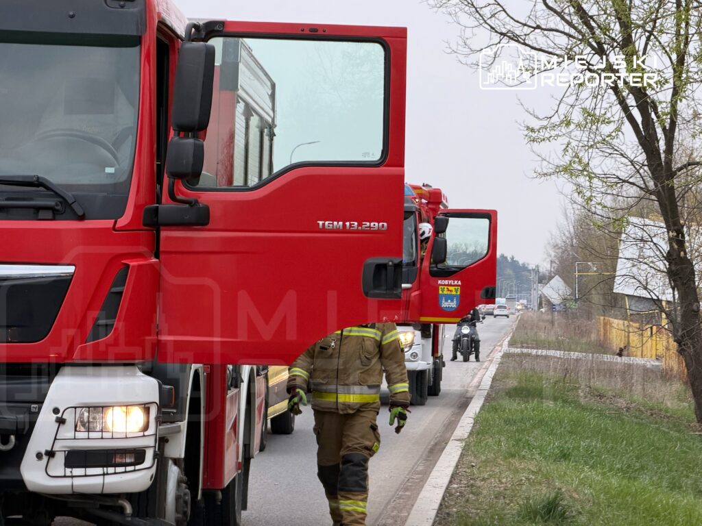 Czerwony wóz strażacki z otwartymi drzwiami, strażak w mundurze wyłania się z kabiny, droga w tle.