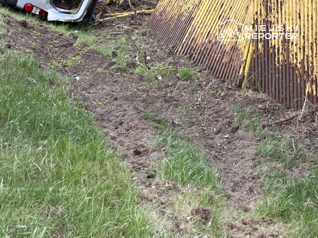 Świeżo wykopana ziemia z widocznymi śladami po oponach w trawie obok metalowego ogrodzenia.