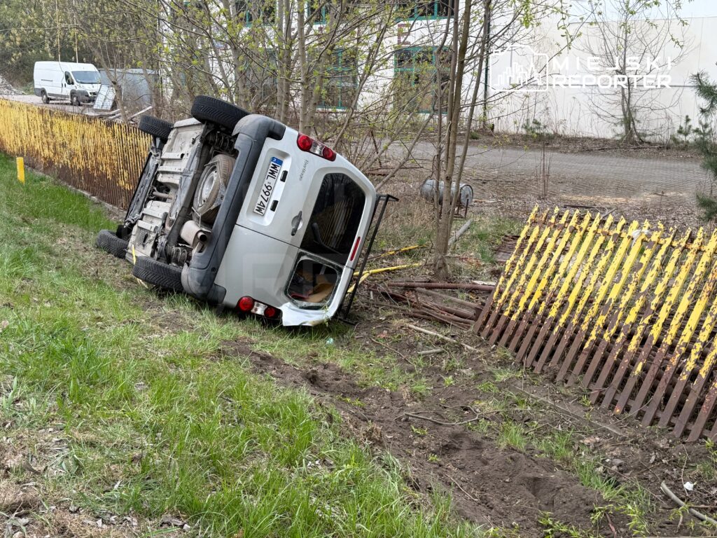 Samochód dostawczy leży na boku w trawie obok uszkodzonego ogrodzenia w zalesionym terenie.