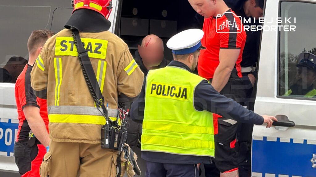 Ratownicy medyczni oraz policjanci pomagają poszkodowanemu mężczyźnie wsiadać do radiowozu.