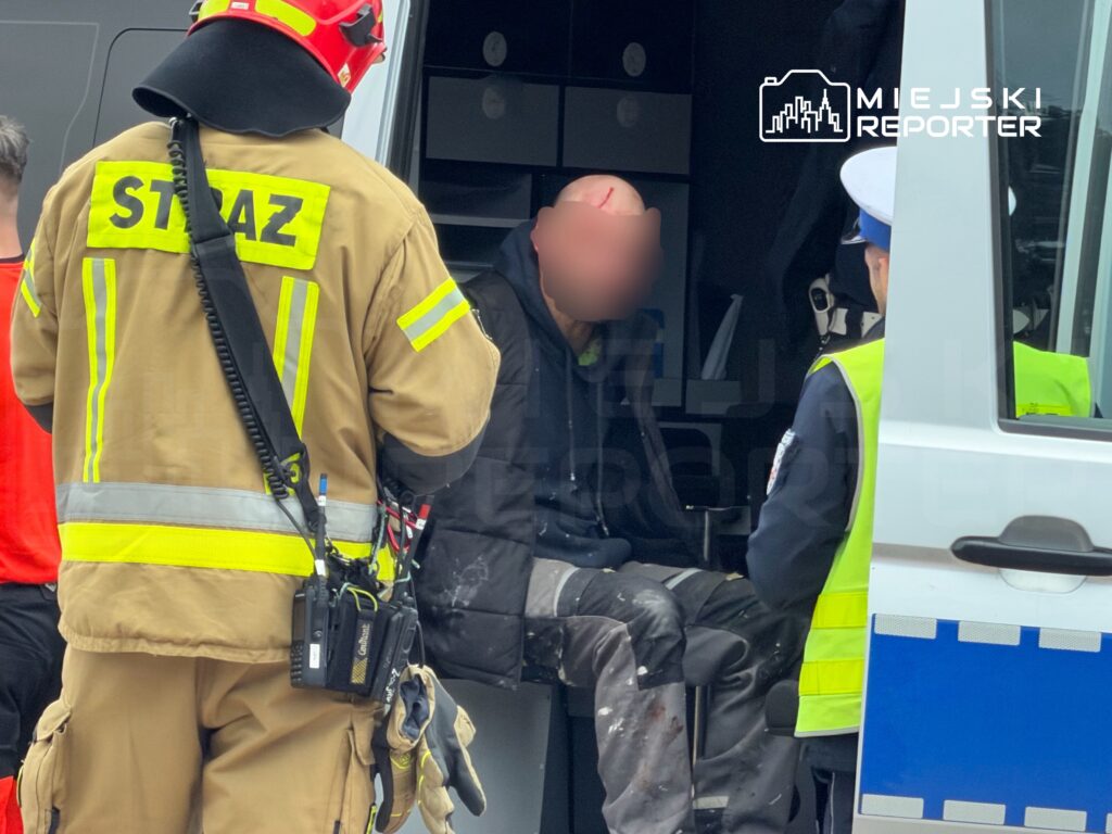 Strażak w mundurze rozmawia z mężczyzną w ubraniu roboczym siedzącym w karetce, z widoczną raną na głowie.