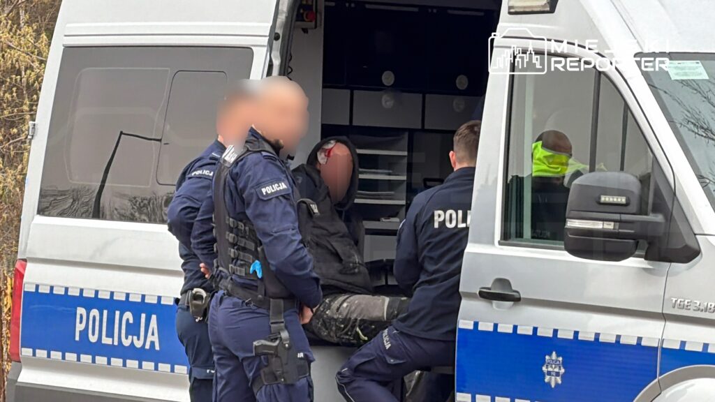 Czterech policjantów udziela pomocy mężczyźnie z raną na głowie, siedzącemu w policyjnym samochodzie.