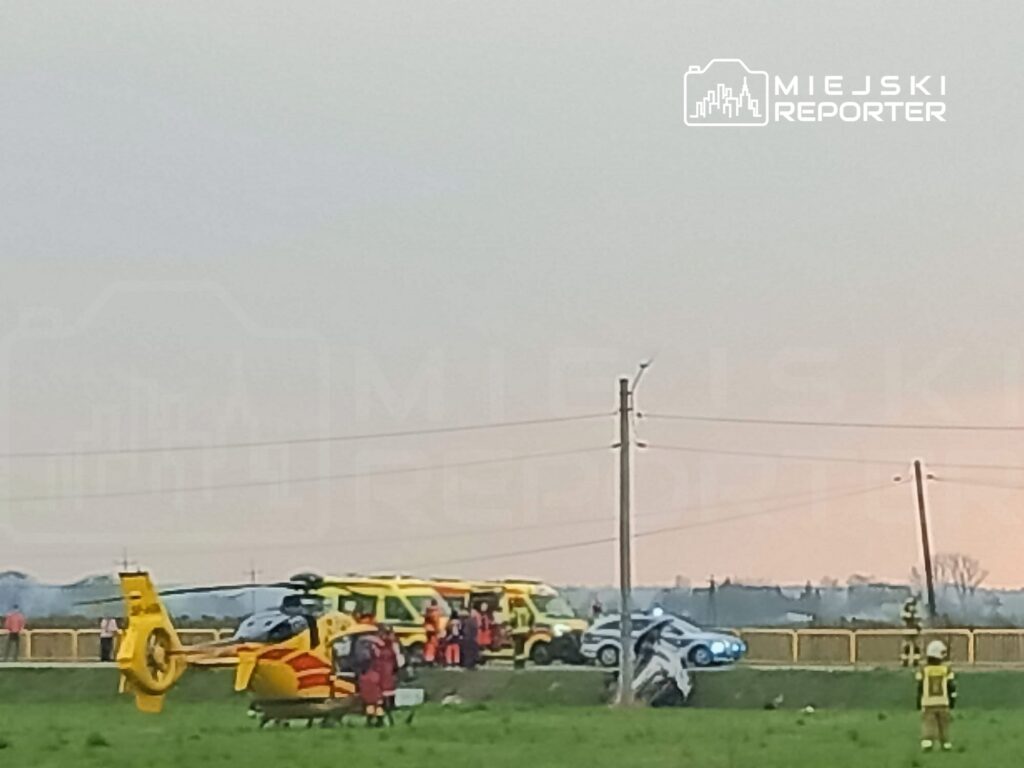 Na łące lądował śmigłowiec ratunkowy, a obok niego stoją pojazdy służb ratunkowych oraz ratownicy w odzieży ochronnej.