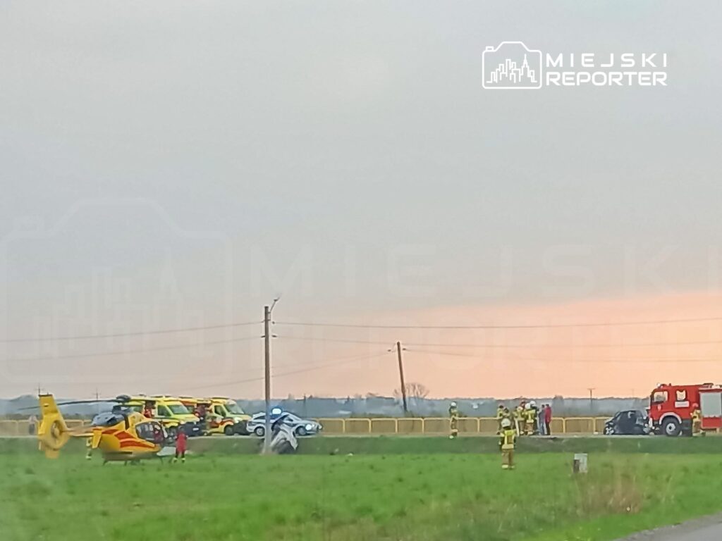 Na polu lądowanie śmigłowca ratunkowego, obok zespół ratowników i wozów strażackich przy uszkodzonym pojeździe.