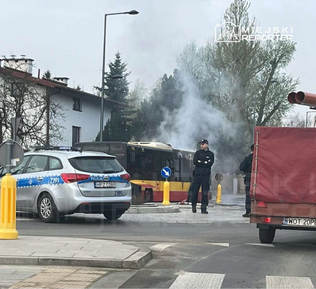 Policyjny radiowóz oraz strażak obserwują dym wydobywający się z autobusu na ulicy w otoczeniu budynków mieszkalnych.