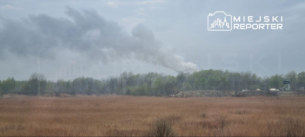Gęsty dym unoszący się z przemysłowego terenu w otoczeniu drzew i łąk.