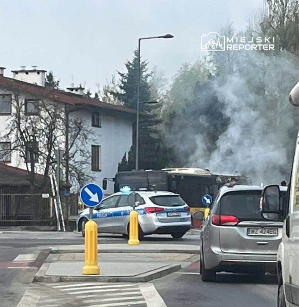 Policyjny radiowóz z włączonymi sygnałami świetlnymi stoi przy skrzyżowaniu, obok dymiącego pojazdu.