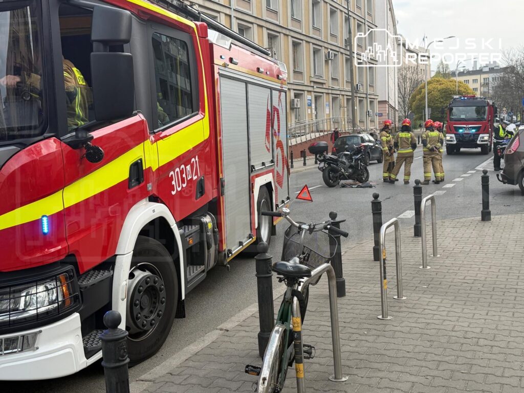 Strażacy w mundurach prowadzą działania ratunkowe przy wypadku motocyklowym na ulicy, obok zaparkowanej roweru.