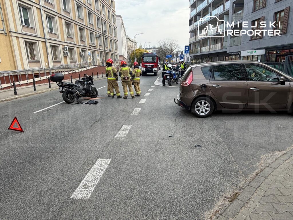 Na jezdni leży przewrócony motocykl, obok grupka strażaków w mundurach, a w tle widać zaparkowane samochody.