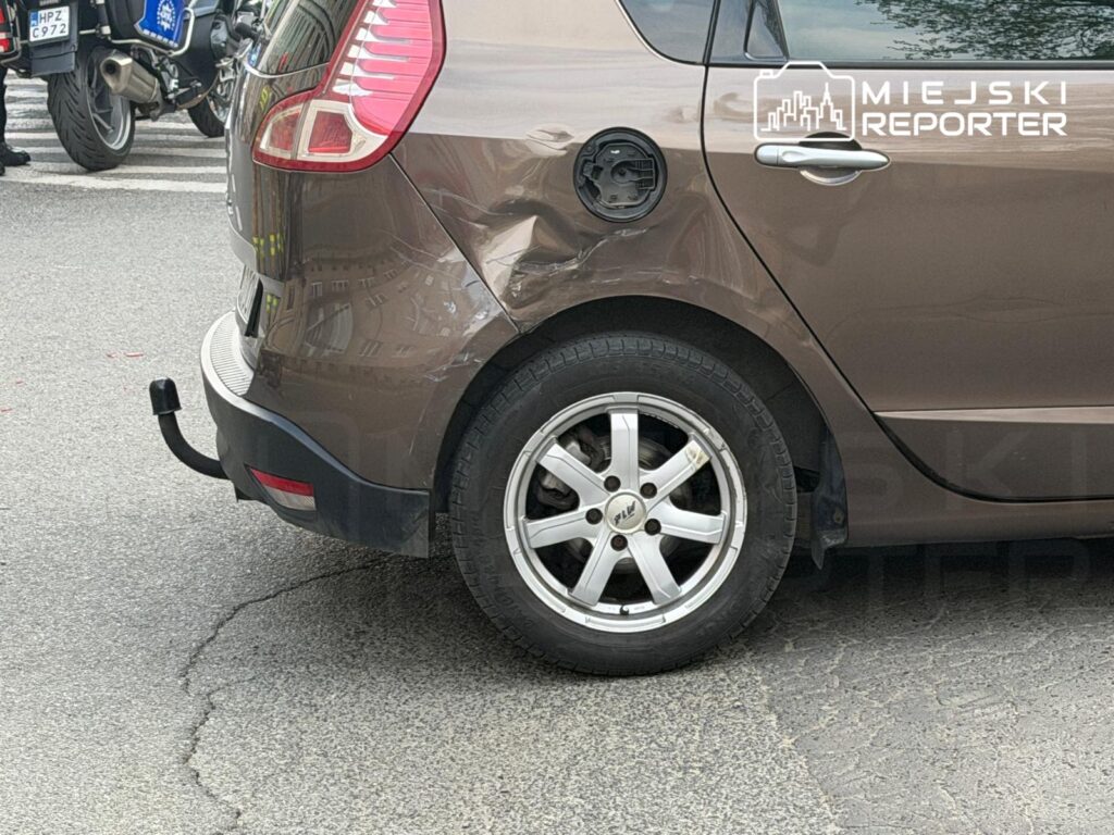 Uszkodzony tył brązowego samochodu osobowego zaparkowanego na ulicy. W tle widać motocykl oraz przejście dla pieszych.
