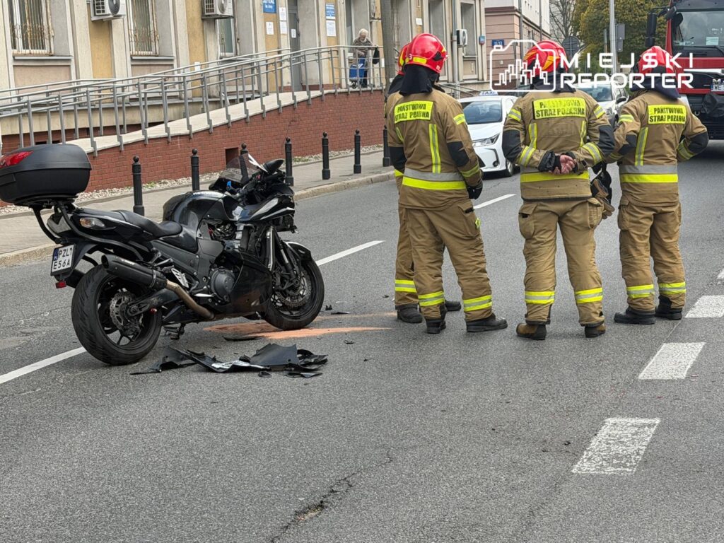 Motocykl leży na jezdni, a obok czterech strażaków w mundurach omawia sytuację po wypadku.