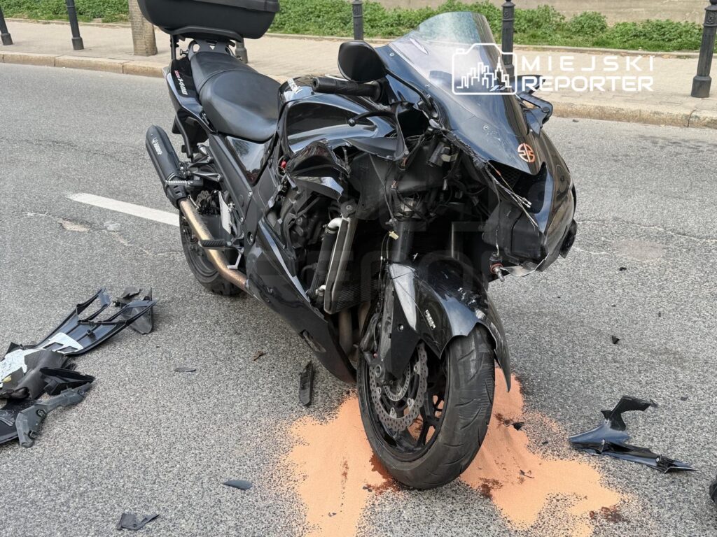 Czarny motocykl leży na jezdni, z uszkodzoną owiewką i odłamkami plastiku wokół, obok widoczne ślady płynów.