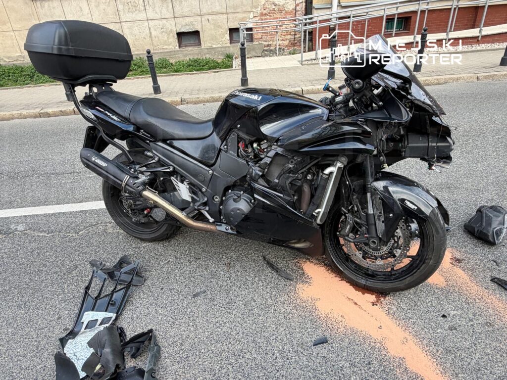 Uszkodzony czarny motocykl leży na jezdni, obok rozbite elementy oraz plama płynu. W tle widoczna ulica z budynkami.