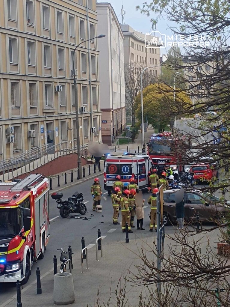 Grupa strażaków w hełmach i mundurach udziela pomocy poszkodowanym na miejscu wypadku drogowego, obok przewróconego motocykla.