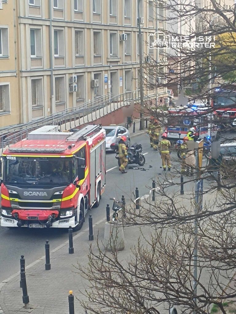 Czerwony wóz strażacki parkuje na ulicy, a strażacy prowadzą działania wokół motocykla i pojazdów zaparkowanych w pobliżu.