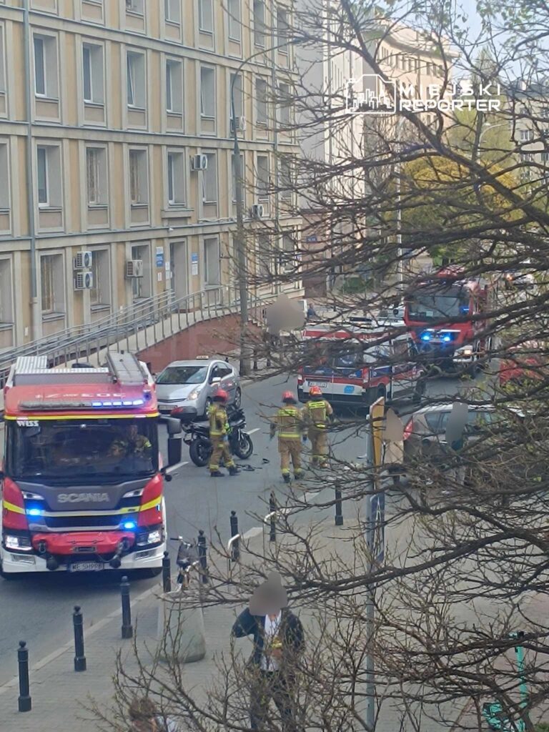 Strażacy w uniformach pracują przy wypadku drogowym, obok przewróconego motocykla na ruchliwej ulicy.