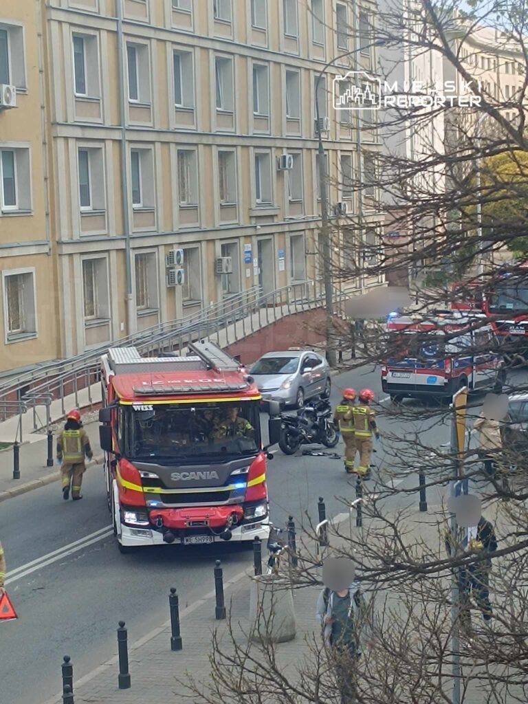 Strażacy w mundurach pracują przy wypadku drogowym, obok przewrócone motocykle i strażacki wóz na ulicy.