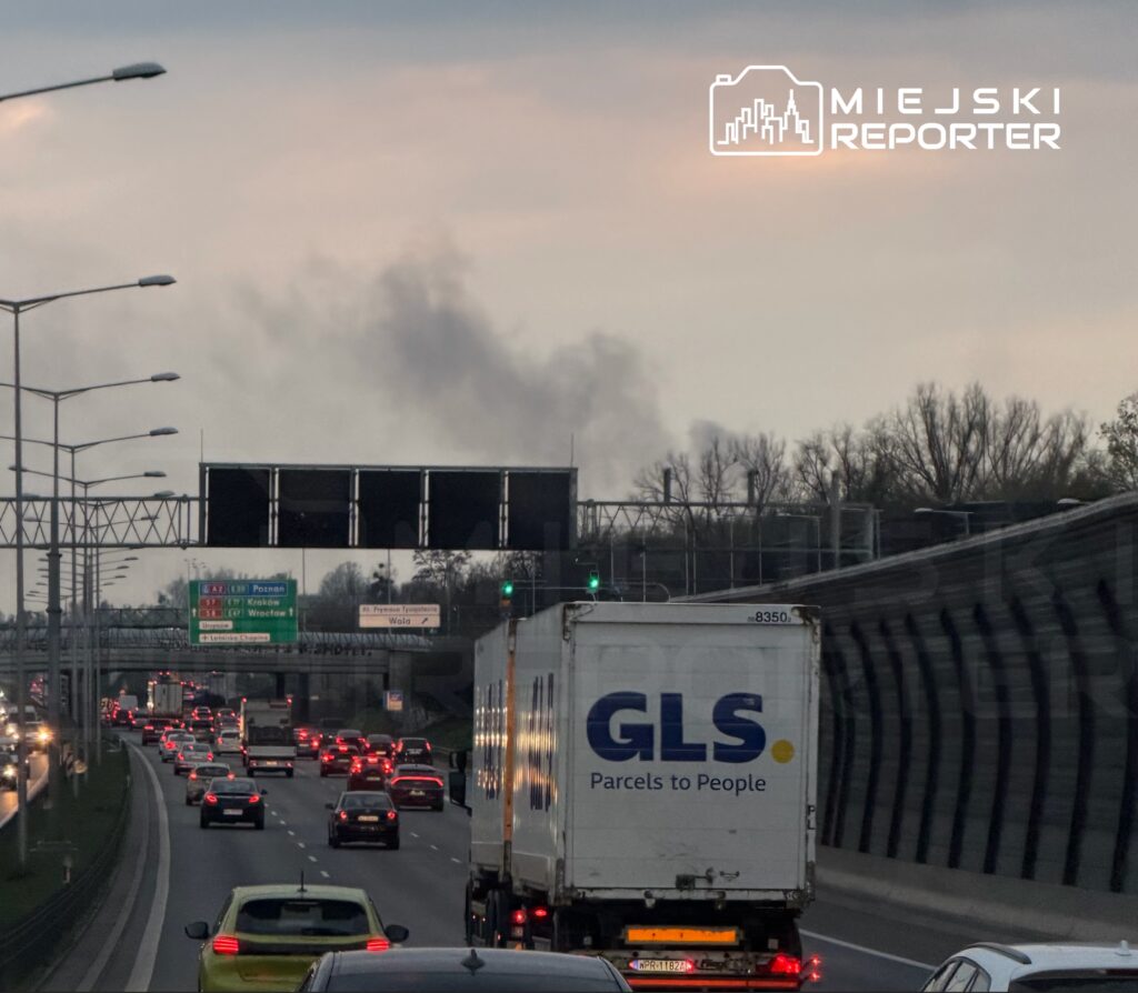 Ruchliwa autostrada z samochodami osobowymi i ciężarówką GLS, w tle widoczne dymiące chmury.