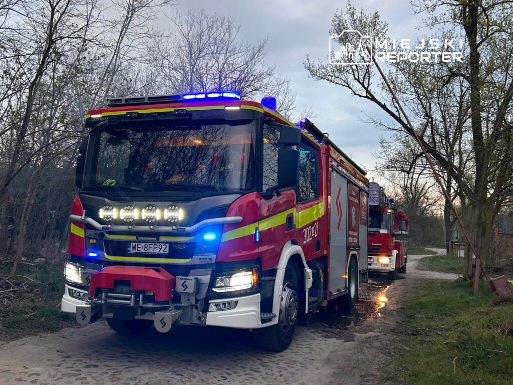 Czerwony wóz strażacki z włączonymi sygnalizatorami świetlnymi stoi na wąskiej drodze otoczonej drzewami.