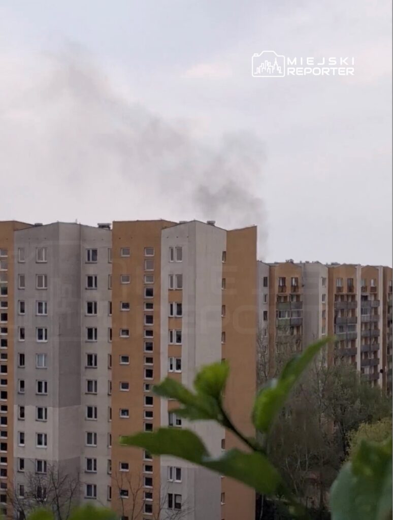 Z dala widoczne bloki mieszkalne z dymem unoszącym się z jednego z budynków. W tle niebo z chmurami.