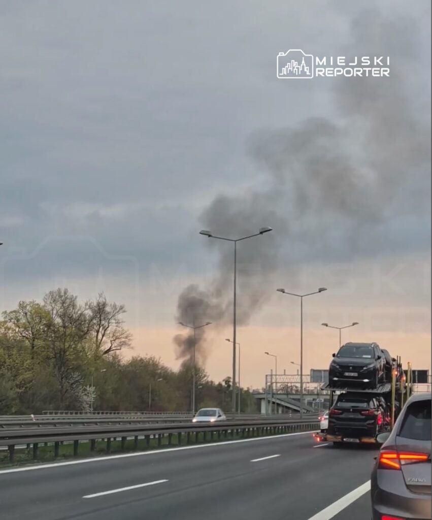 Dym unoszący się nad drogą, w tle pojazdy na autotransporcie, w otoczeniu zieleni i lamp ulicznych.