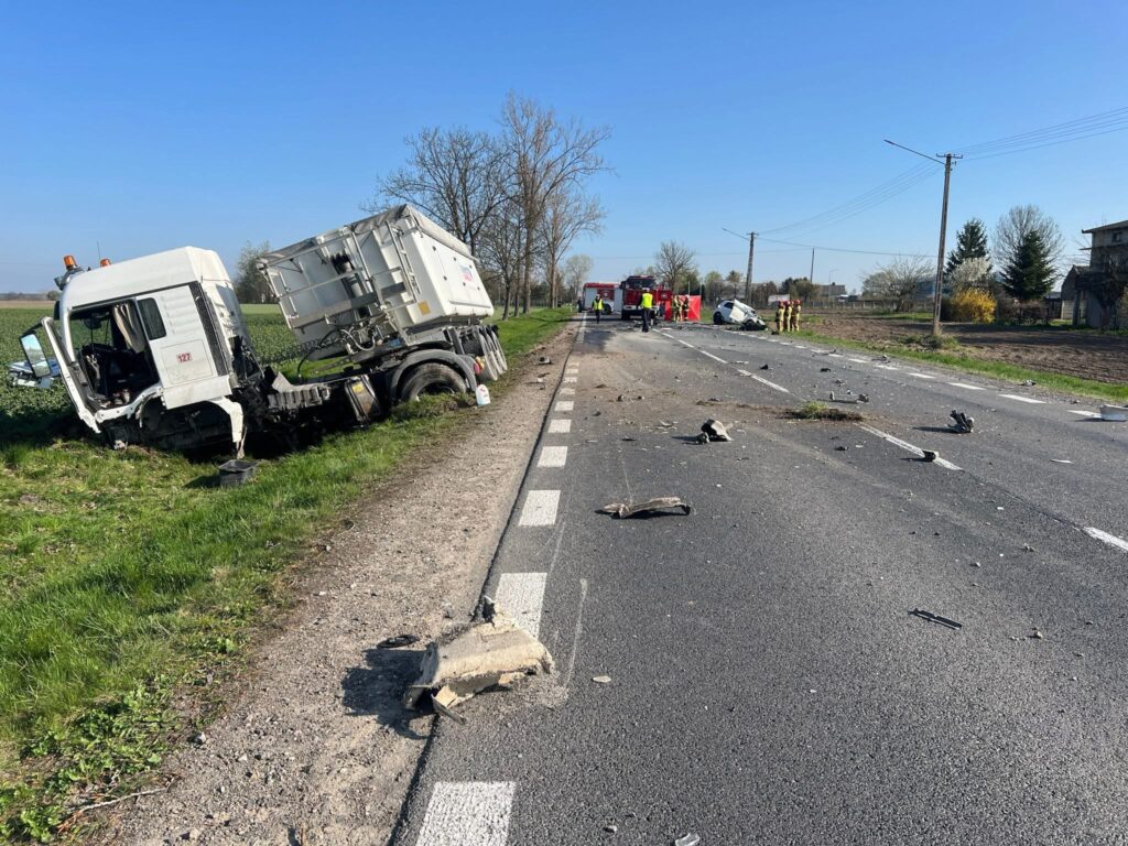 Uszkodzony ciężarówka leży w rowie obok drogi, a w tle pracują służby ratunkowe przy wypadku drogowym.